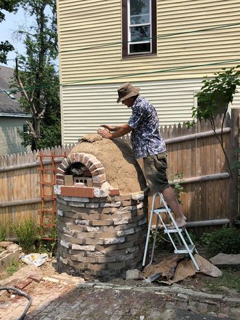 A person stands on a short ladder, working on building the upper portion of an outdoor oven