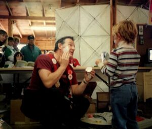 Clown putting on makeup while a child holds a mirror