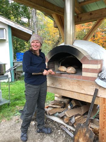 A person stands with a wood paddle, retrieving freshly baked bread loaves from an outdoor oven