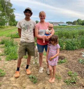 a man in a white tshirt, shorts and orange shoes stands in a farm field with a woman in a pink and red shirt; she holds a baby and young girl in a floral dress leans against her leg