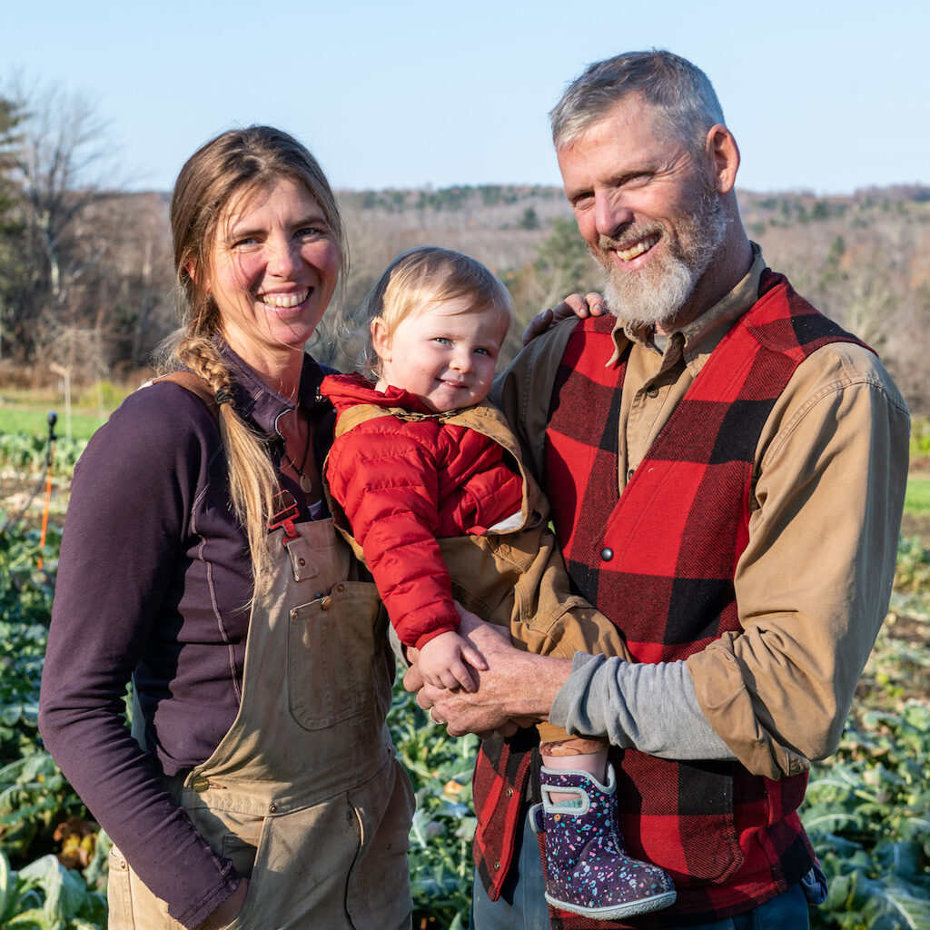 Two farmers smiling in a crop field, holding a baby