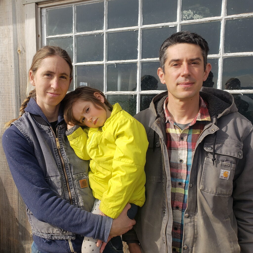 Two farmers holding their child, standing in front of a window