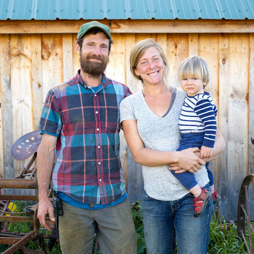 Two farmers smiling in front of a barn, holding their child