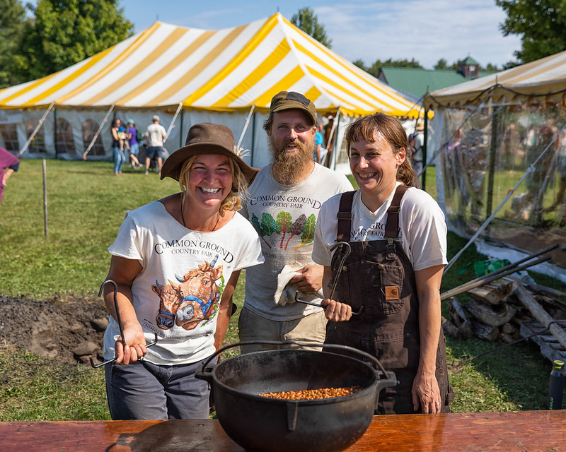 Three smiling volunteers pose in front of a bean pot at the Common Ground Country Fair.