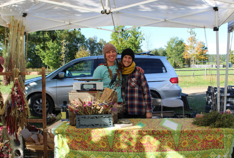 Two farmers smiling at a farm stall filled with amaranth.