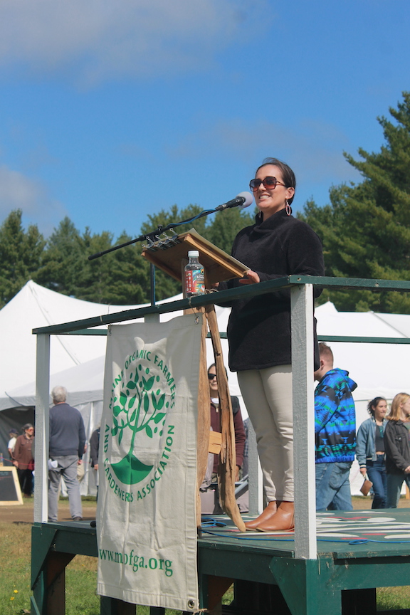 Maulian Dana delivers a keynote speech from a wooden podium adorned with a canvas banner, which bears the green tree logo of the Maine Organic Farmers and Gardeners Association.