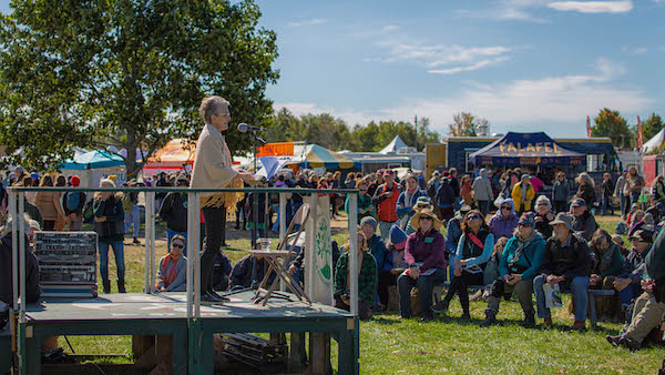Frances Moore Lappe delivers a keynote speech from a wooden podium before a crowd of fairgoers seated on hay bales and a green lawn.