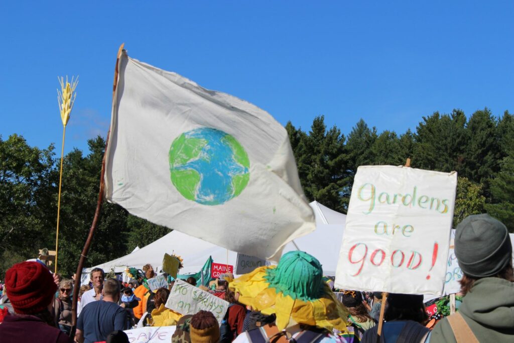 A flag with a picture of the earth and a sign that says "Gardens are Good" in a crowd of people.