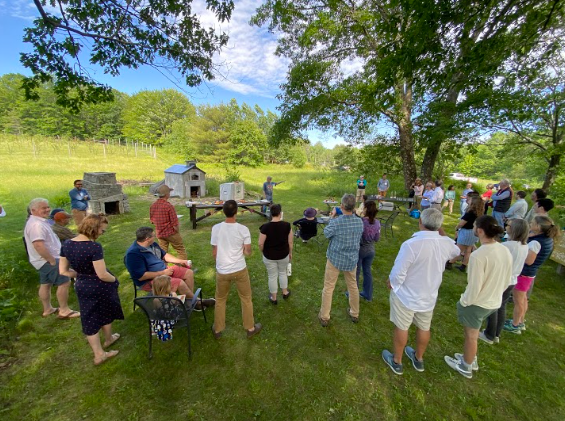 People stand in a scattered circle around a speaker at an outdoor event.