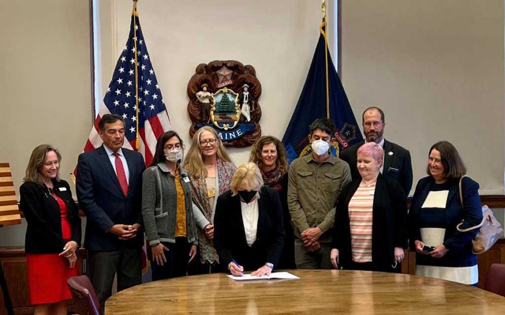 Governor Mills signs a bill into law in Augusta, while policy workers watch.