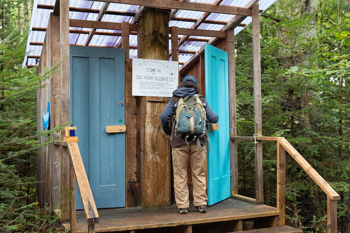 A fairgoer wearing a ballcap and a backpack opens the blue door to an outhouse