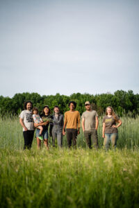 Six farmers stand smiling in a field; one holds a small child