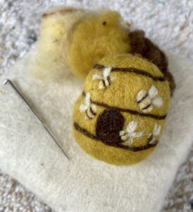 A felted beehive sits on a felt pad next to a needle.