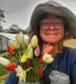 A farmer in a hat and glasses poses with a bouquet of daffodils and tulips.