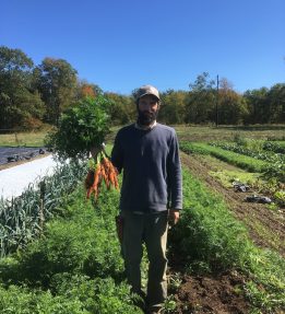 Farmer Jeff Benton stand in the pathway between two lush beds of carrots. He is holding a bunch of bright orange carrots by their green tops.