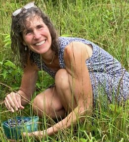 MOFGA board member, Jo D. Saffeir kneeling in tall grass and holding a pint of blueberries.