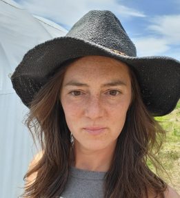 A person wearing a black wide-brimmed sun hat takes a selfie in a farm field.