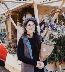 farmer in flower crown smiling holding a bouquet in front of farm stand