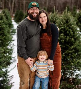 Two farmers and a toddler stand, smiling, in front of a Christmas tree field.